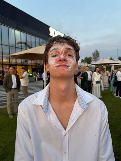 This is a medium shot, vertically oriented, candid photograph of a young man with white cream or frosting smeared across his face and hair. He is wearing a white button-down shirt, partially unbuttoned. He is looking directly at the camera with a slight smile, his eyes wide. The background suggests an outdoor gathering with other people visible, some appearing to be dressed formally. Behind them, there is a modern building with large glass windows and an illuminated sign that partially reads "MKR". Tents and umbrellas are also present, indicating an event or celebration. The grass in the foreground suggests a park or outdoor venue. The lighting appears to be from late afternoon or early evening, with a soft, natural light. The scene conveys a sense of playful celebration and camaraderie. The location is Khotianivka, Ukraine.