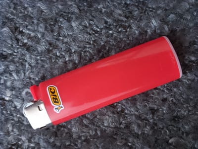 A red BIC lighter lies diagonally on a dark gray, fuzzy surface. The lighter is positioned with its metal end to the left and the ignition mechanism pointing upwards. The brand logo "BIC" is visible on the side of the lighter in yellow and black lettering with a small graphic of a stick figure. The fuzzy surface appears to be a fabric material, possibly a rug or blanket, with a high pile. The lighting is even, and the image is taken from a top-down perspective. There are no people or actions depicted in the image. The scene does not provide any information that can be used to determine its location in Kropyvnytskyi, Ukraine.