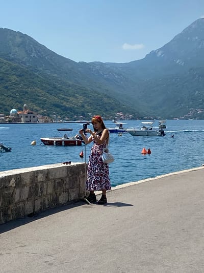 A woman wearing a red-and-white patterned dress and a red headband stands on a paved walkway next to a stone wall, holding up a smartphone to take a selfie. She has dark hair and is looking at the phone. Her purse is slung over her shoulder. Behind her, the blue water of a bay is dotted with several boats. In the distance, a red-and-white boat is moored, and beyond that, a domed building with a blue roof is visible on a small island. Steep, green, tree-covered hills rise behind the bay, under a clear blue sky. The scene is illuminated by bright sunlight, casting shadows on the walkway.