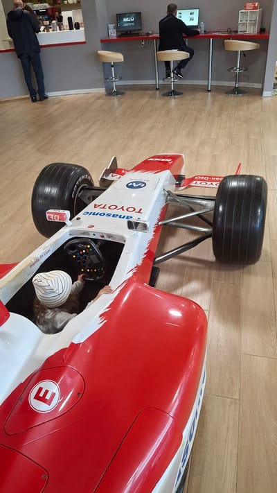 The image depicts the interior of a public space with a red and white Formula 1 racing car as the central subject. The car's cockpit is occupied by a child wearing a striped hat, who is holding the steering wheel. The car has "TOYOTA" and "Panasonic" branding. In the background, two men are seated at a red counter with computers. One man is facing a monitor, while the other is looking away from the camera. There are bar stools around the counter. The flooring is made of light-colored wood planks. The location is identified as Novogorelovo, Russia.
