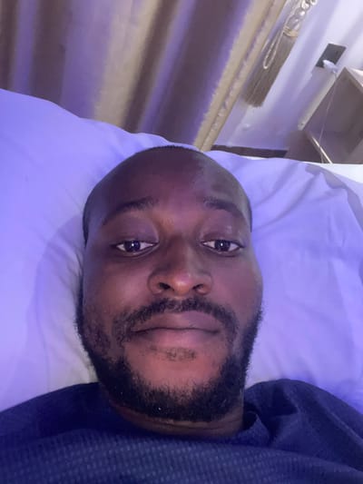 A male individual is depicted in a close-up, top-down selfie, lying in what appears to be a bed. The scene is illuminated primarily by strong purple and blue ambient light, casting distinct hues over the environment. The individual has dark skin, a shaved head or very short dark hair, and a full, dark beard and mustache. His dark eyes are open, looking directly into the camera, exhibiting reflections. He is wearing a dark blue, textured long-sleeved garment, visible across his chest and shoulders. His head rests on a white pillow, which, along with white bedding material, is visible beneath and to the sides of his head, all significantly tinted by the purple light. In the background, to the upper right, a wall is visible, featuring an electrical outlet with a white cable plugged in and trailing downwards. Below the outlet, a portion of a light brown wooden shelf or furniture piece is present. To the left and slightly behind, a textured curtain or wall covering, also infused with the purple light, is discernible. A decorative tasseled cord hangs from the wall near the curtain. The specific location is Galadimawa, Nigeria.