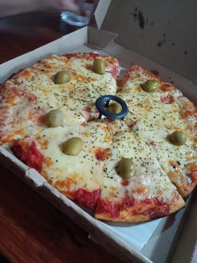 A pizza with cheese, tomato sauce, and green olives in a cardboard pizza box is on a wooden surface. The pizza is cut into slices, with a black metal ring in the middle. The location is in El Talar, Argentina, based on coordinates (-34.473, -58.639). The pizza is the subject of the image. It is in the center of the image and the entire frame focuses on it. The background is a blurry wooden surface. The pizza appears to be fresh and ready to be eaten. The pizza is slightly tilted to the right and the slices are slightly overlapping. There is white paper on the bottom of the box underneath the pizza. The pizza box is open and the lid is slightly folded back behind the pizza. The ring in the center is not touching the pizza.