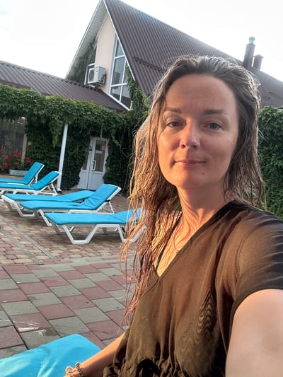 A woman with wet, dark brown hair and fair skin is taking a selfie outdoors. She is wearing a sheer black t-shirt. Her face is turned towards the camera, with a neutral expression. The background shows a building with a brown, gabled roof covered in green vines. Several blue lounge chairs are arranged on a brick-patterned patio. An air conditioning unit is mounted on the side of the building. The scene suggests a sunny day, possibly at a resort or outdoor recreational area.