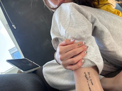 A person is sitting indoors, wearing a grey sweatshirt. They are holding their left hand, which has manicured nails, over their right wrist. The right wrist has a tattoo consisting of numbers and symbols. A tablet device rests on the person's lap to the left. A yellow garment is visible near the person's right shoulder. The location is indiscernible in the image; therefore, the city of Hull, United Kingdom, cannot be confirmed.