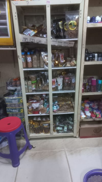 This is a close-up, eye-level shot of a tall, narrow shelving unit filled with various food products and toiletries. The shelving unit is light colored, possibly wood or laminate, and is divided into multiple shelves. The scene appears to be indoors, possibly a small shop or a storeroom.

On the shelves, there are many packaged snacks, nuts, and dried fruits. Some prominent items include clear plastic bags of nuts with colorful labels, larger bags of what appear to be crispy snacks, and smaller foil-wrapped packages. There are also bottles and jars of various sizes. On the right side of the shelving unit, on the upper shelves, there are several small bottles, likely toiletries such as roll-on deodorants or perfumes, with visible branding like "Nivea" and "Axe". Below these, on the lower shelves of the right section, are more toiletries and small containers, some brightly colored.

To the left of the shelving unit, stacked boxes are visible, one with "Fayrouz" branding, suggesting beverages. A bright purple stool with a red seat is positioned in the foreground, partially obscuring the bottom left of the shelving unit. The floor is tiled with a light-colored, possibly ceramic or linoleum material. The lighting is moderate, and the overall impression is of a well-stocked, albeit slightly cluttered, retail space. The location is described as Rayfield, Nigeria.