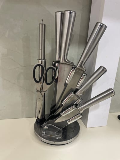 The image shows a knife block with several knives and a pair of kitchen shears. The knife block is made of stainless steel and has a round base. The knives have stainless steel handles and blades. The kitchen shears also have stainless steel handles and blades, with black rubber grips on the handles. The knife block is placed on a white countertop. The background is a light-colored tiled wall. There are no people visible in the image. The scene is indoors, likely in a kitchen. The location of Slobozia-Dușca, Moldova, is not evident from the image.