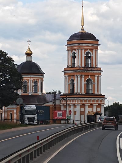 This is an eye-level, medium shot of an Orthodox church with a bell tower. The church has an orange facade with white columns and arches, and a dark dome topped with a golden cross. The bell tower is also orange and white, with multiple levels of arches and columns, and a tall, slender golden spire. In front of the church, a large white truck with a blue shipping container is parked. A red sign with Russian text and a QR code is visible on the side of the building. A road with a metal guardrail curves in the foreground, with two cars visible on the road. The sky is cloudy and overcast. The location is the city of Khotilovo, Russia.