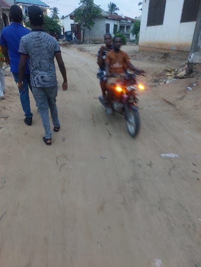The image depicts an unpaved dirt road in Mwandege, Tanzania, under dim light suggesting dusk or early evening conditions. In the left foreground, three individuals walk away from the camera. The closest wears a horizontal black and white striped short-sleeved shirt, grey pants, and black sandals. Ahead of him, another individual wears a blue short-sleeved shirt and blue jeans. A third person is partially visible on the far left. On the right side of the road, a motorcycle with two occupants is in motion, blurred from motion, and approaches the camera, displaying two illuminated forward lights. The driver wears an orange-brown short-sleeved shirt, with a passenger seated directly behind. Residential buildings, including a prominent white structure and another with a light blue roof, line the background. Scattered debris and sparse vegetation are visible along the road's edges.