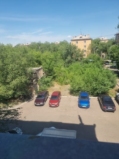 A high-angle, full shot captures a city scene under a clear blue sky with sparse white clouds. In the foreground, the top of a blue-tinted structure, possibly a balcony or roof, is visible, casting a large shadow on the ground below. The middle ground features a parking lot with five cars. From left to right, a dark red sedan, a red hatchback, a bright blue hatchback, and a dark blue sedan are parked. The blue hatchback has a license plate ending in "09". The dark blue sedan's license plate ends in "U". To the far right, a silver car is partially visible. Behind the parking lot, dense green trees form a thick canopy, obscuring much of the lower buildings. To the left, a crumbling stone structure is partially covered by greenery. In the background, several apartment buildings with light-colored facades and dark roofs are visible, interspersed with more trees. Streetlights and power lines are also present. The image appears to be taken in the city of Saran, Kazakhstan, given the presence of Cyrillic script on the license plates.