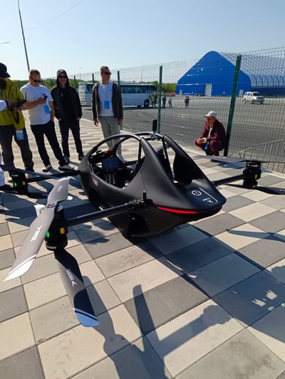 In Korsakov, Russia, on a sunny day with a clear blue sky, a black drone with a cockpit and four rotors sits on a tiled walkway. The drone is the primary subject of the image, with its sleek, aerodynamic design and visible propellers. Several individuals are gathered around the drone, observing it. To the left of the drone, a group of four men are standing. One man on the far left, wearing a yellow jacket and a dark cap, holds a remote control. Next to him, a man in a white t-shirt and sunglasses looks at his phone. A man in a black hoodie and sunglasses stands in the middle, with his hands in his pockets. The fourth man, also in a white t-shirt and sunglasses, is dressed in a dark jacket and light-colored pants. Further to the right, a person in a maroon jacket and cap is crouched down near a fence, appearing to observe the drone from a distance. In the background, a large blue arched building is visible, along with a chain-link fence and parked vehicles, suggesting an outdoor event or exhibition. The overall scene depicts a technological display with onlookers.