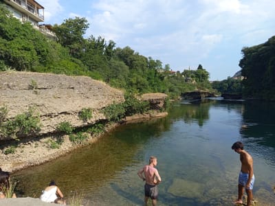 The image captures a serene riverside scene under a bright, partly cloudy sky. The river, with its clear, shallow waters, flows gently, reflecting the lush green trees that line its banks and the surrounding rugged cliffs. On the left bank, a multi-story building, possibly a residential complex, is nestled amongst the dense foliage, its balconies overlooking the water. In the foreground, a few individuals are enjoying the riverside. On the far left, a person with dark hair is partially visible, and next to them, another individual in a white top is bending down towards the water. Further into the river, a man with blonde hair and visible tattoos stands with his hands on his hips, facing away from the camera. To the right of him, another man, wearing blue patterned shorts, stands looking down into the water. The overall impression is one of a tranquil summer day, with people relaxing by the river in a natural, verdant setting. This scene is likely near the Kravice waterfalls or a similar riverine area close to the city of Mostar, Bosnia and Herzegovina, known for its natural beauty and the Neretva River.