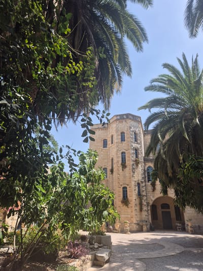 In Abu Gosh, Israel, a sandstone building with multiple stories and arched windows is partially obscured by lush green foliage and palm trees. The building's facade features an octagonal tower on the left and a central arched entrance on the right. The courtyard in front of the building is paved with gravel and stone pathways, with potted plants and decorative stones scattered around. The sky is clear and blue, suggesting a sunny day. The overall impression is one of a tranquil and historic location, possibly a monastery or a historical estate, surrounded by abundant vegetation.