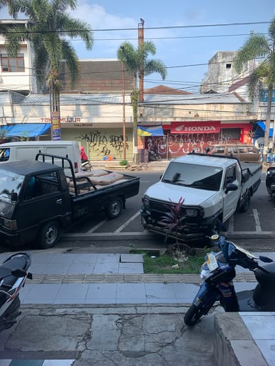 In Sumedang, Indonesia, a street scene unfolds under a bright sky, likely in the morning given the clear lighting. The foreground is dominated by a cracked concrete sidewalk and a section of grey tiled pavement. To the right, a blue and black Yamaha scooter is parked, angled towards the street, with a license plate reading "E 4263 CZ".
The street itself is paved and marked with white lines indicating parking spaces. A white pickup truck is parked in one of these spaces. To the left, a black pickup truck is also parked, its bed filled with numerous sacks of what appear to be cement or similar building material. A white van is parked behind the black pickup. A person wearing a pink jacket and a helmet is seated on a motorcycle to the left of the white van, looking to the right.
Behind the vehicles, a row of buildings lines the street. The buildings have awnings and storefronts. One storefront displays a large red "HONDA" logo. Another storefront has signs with "TOKO MUJUR" and "BELI TV 100 UANG KEMBALI". Graffiti covers the lower sections of some of these buildings. Palm trees are visible in the background, with their fronds reaching towards the sky. Power lines crisscross the scene overhead. The overall impression is one of an active commercial street in Indonesia.