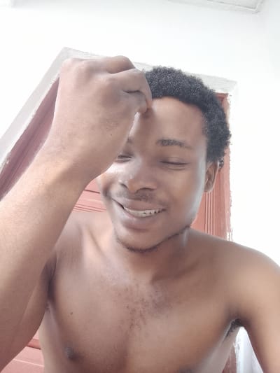 The media file is a close-up, eye-level selfie taken by a young Black male. He is shirtless and appears to be outdoors, with a white wall and a reddish-brown wooden door visible in the background, suggesting a domestic setting in Orodo, Nigeria. The lighting is bright, indicating daytime.

The subject has dark, curly hair and is wearing a slight smile, with his eyes closed. His right hand is raised to his forehead, fingers lightly touching his hairline. His skin is smooth, and he has a light stubble on his chin and upper lip. His chest and upper arms are visible, showing a clear complexion.

The overall mood of the image is relaxed and content. There are no visible text elements in the media file.