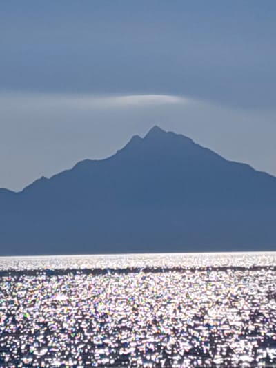 This is a vertical, medium-shot, outdoor photograph of a mountain range silhouetted against a hazy sky over a shimmering body of water. The mountain range is dark blue and appears to be a single, continuous formation with jagged peaks. A thin, lighter-colored horizontal cloud formation is visible directly above the mountain range. The water in the foreground is bright and reflects the sunlight, creating a dazzling, speckled effect with iridescent colors. The overall atmosphere is serene and natural, possibly depicting Mount Athos in Halkidiki, Greece, a location known for its dramatic mountain scenery overlooking the Aegean Sea.