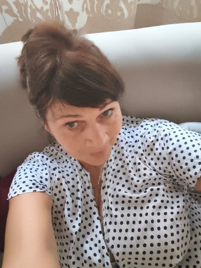 A woman with dark brown hair pulled up is looking at the camera. She is wearing a white shirt with black polka dots. The background is a light-colored couch with a patterned wallpaper visible behind it. The image appears to be a selfie taken indoors. There is no information available to determine the location in Livorno, Italy.