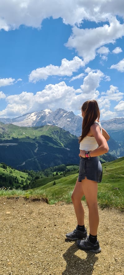 In a stunning mountainous landscape, a person is captured from the waist down, performing a push-up. The scene is set on a gravel path with patches of green grass on the left, indicating an outdoor environment. The background is a breathtaking panorama of the Dolomites in Val Gardena, Italy, featuring snow-capped peaks, lush green slopes, and a winding road visible in the distance. The sky is a vibrant blue, dotted with fluffy white clouds, suggesting fair weather.
The subject is wearing a white tank top, dark shorts, black socks, and athletic shoes, suggesting they are engaged in some form of physical activity or hiking. The sun is casting a noticeable shadow on the ground, implying it's daytime, likely midday given the brightness. The person's posture suggests focus and exertion during the push-up. The overall impression is one of strength and determination amidst a picturesque and awe-inspiring natural setting. There is no visible text in the image.