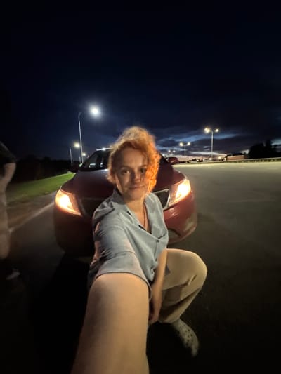 A woman with bright orange curly hair is taking a selfie at night next to a red car. She is wearing a light blue button-up shirt and tan sweatpants. Her left arm is extended towards the camera, obscuring part of her face and body. The car's headlights are on, illuminating the road and the surrounding area. In the background, there are streetlights visible along a highway. The sky is dark with some clouds and the twilight colors of sunset. The scene appears to be an outdoor location, possibly a roadside or parking area, in or near Alexeyevskoye, Russia.