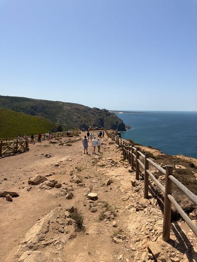 The image captures a scenic view of the coastline near Colares, Portugal, specifically at Cabo da Roca, the westernmost point of mainland Europe. A well-worn dirt path, lined with wooden fencing, winds along the edge of a cliff overlooking the vast, deep blue Atlantic Ocean. Several tourists, dressed in casual summer attire, are walking along the path, some in small groups and others individually. The terrain is rocky and uneven, with scattered stones and sparse vegetation clinging to the dry, earthy soil. Lush green hills rise in the background on the left side of the image, contrasting with the arid cliff face. The sky is a clear, bright blue, indicative of a sunny day.