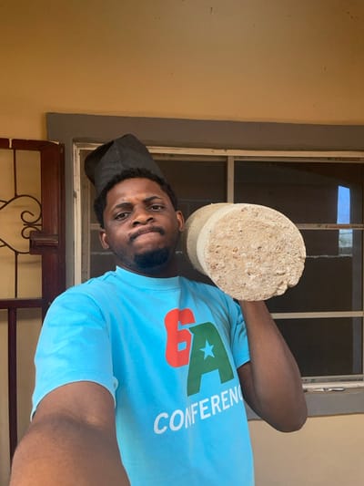A male individual, presumed to be in Ube, Nigeria, is depicted in a chest-up shot. He has a dark complexion, short dark hair, and a trimmed beard, wearing a black, patterned traditional cap. His facial expression is firm, with direct eye contact towards the camera. He is clad in a light blue short-sleeved t-shirt displaying a logo featuring a red number '6', a green letter 'A' with a white star, and the word "CONFERENCE" in white text below. The individual holds a light beige, rough-textured cylindrical object, consistent with a concrete test cylinder, on his left shoulder, supported by his left arm. The background consists of a light beige wall. To the left, a portion of a dark brown wooden door frame with carved details is visible. Centrally located behind the individual is a window with visible horizontal white or light-colored frame divisions, possibly security bars, and a thin, metallic-colored frame. The lighting indicates a daytime setting.