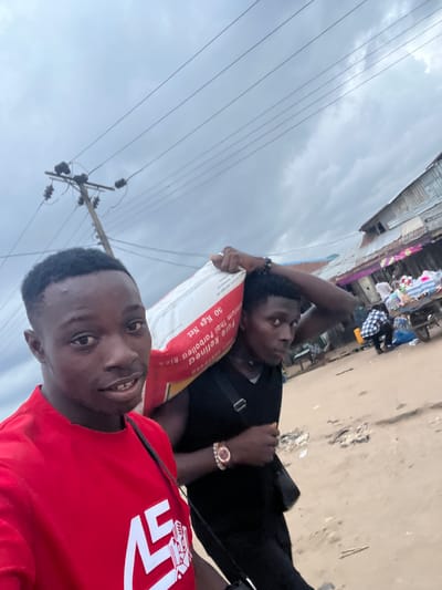 In Oko Amakom, Nigeria, two young men walk down a sandy street under a cloudy sky. The man in the foreground, wearing a red t-shirt with a white graphic, smiles at the camera. Behind him, a second man carries a large, white bag of "Pure Refined Thai parboiled Rice 50 Kgs Net" on his shoulder. The background shows a row of tin-roofed buildings, possibly market stalls, with people and a cart visible. A utility pole with electrical wires stands to the left. The ground is composed of light brown sand and scattered debris.