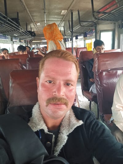 An interior shot, likely taken on a train, shows a fair-skinned male with reddish hair and a prominent mustache, dressed in a dark jacket with a fur-lined collar. He is looking directly into the camera. Behind him are rows of brown, leather-like seats occupied by other passengers of varying ethnicities. Some passengers are looking out the windows, some are using their phones. Overhead luggage racks are visible on the right side of the frame. The background features advertisements in Hindi, suggesting the scene is located in India, likely in Orai, based on the prompt.