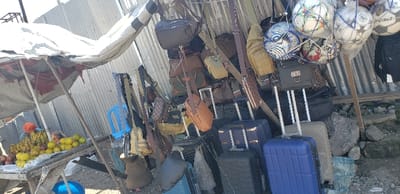 The image shows a street vendor selling luggage and bags in Rubavu, Rwanda (latitude: -1.668, longitude: 29.268). There are a variety of suitcases, duffel bags, and backpacks on display, all arranged in a row against a corrugated metal wall. The vendor is standing behind the display, partially obscured from view. A small, white canvas awning provides some shade for the items being sold.  The vendor's stall is positioned between a nearby store with a white awning selling fruit and a small building with a corrugated metal facade.  Several soccer balls are also hanging from the awning.  A blue plastic chair is positioned in front of the vendor's stand.  The ground is unpaved and uneven.  The vendor is not interacting with anyone and is likely awaiting customers.  The image is taken at eye-level.