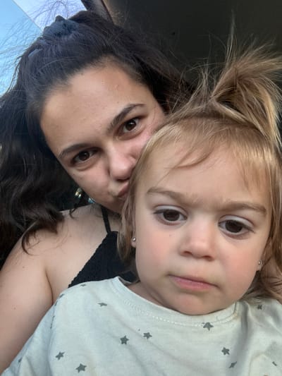 In Oreshak, Bulgaria, a close-up selfie features a woman and a young child. The woman, with dark, wavy hair pulled back, is positioned behind and to the left of the child, her face turned towards the camera with a pout. She wears a black, lacy top. The child, with blonde hair styled in pigtails, is in the foreground, facing the camera with a serious expression. The child's light-colored t-shirt, adorned with grey stars, is visible. The background is dark and out of focus.
