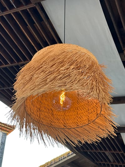The image shows a close-up of a hanging lamp with a lampshade made of natural, straw-like material. The lampshade has a wide, bell shape and is covered in numerous thin, dry strands that extend outwards, creating a fringed effect. A warm-toned Edison-style light bulb with a visible filament is illuminated inside the lampshade, casting a golden glow. The background reveals a wooden slatted ceiling and a white ceiling panel. Part of a red-tiled roof and a grey post are visible on the left side, suggesting an outdoor or semi-outdoor setting. The image does not contain any people or indicate a specific location within the city of Ravda, Bulgaria.