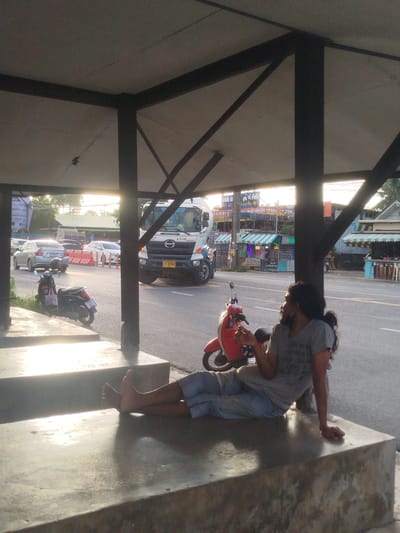 This media file captures a moment in time in Si Sunthon, Thailand, likely during the late afternoon or early evening, judging by the warm, low sunlight casting long shadows. The scene is set under an open-air shelter, possibly a bus stop or a roadside rest area, with a concrete structure in the foreground.
The primary subject is a person with long, dark hair, casually dressed in a t-shirt and shorts, resting on the concrete steps of the shelter. Their bare feet are visible, and they appear to be relaxed, perhaps looking off into the distance. Beside them, a vibrant red scooter is parked. In the background, a busy street is visible with traffic, including a white truck with the license plate "71-8398 A-8749". A black scooter is also parked on the side of the road. Several small businesses with awnings line the street, some displaying signs with Thai text. One sign clearly reads "ซ่อม" (repair) and another mentions "ปิดบริการทุก" (closed every day). The overall atmosphere suggests a typical day in a roadside community.