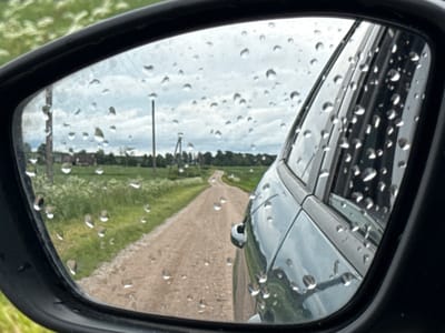 The image shows a view through a car's side mirror in Anglininkai, Lithuania. The mirror is covered in raindrops, which distort the reflection of the environment. The reflection shows a dirt road winding through a rural landscape with green fields and trees. A telephone pole is visible on the left side of the reflection, and in the distance, there appear to be some buildings. The sky is overcast, with clouds contributing to the moody atmosphere. The side of the car, a dark teal color, is also visible in the mirror, with raindrops on its surface. The overall scene suggests a car driving on a wet, country road on a cloudy day in Anglininkai, Lithuania.