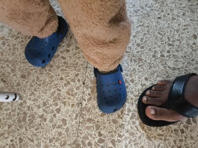 The image shows three feet on a tile floor. Two feet wear blue Crocs. One foot wears a black sandal. The wearer of the Crocs appears to be wearing a brown furry costume. The tiled floor has a speckled pattern in various shades of brown and white. A white object is partially visible in the lower left.