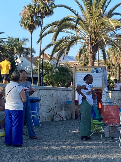 The image shows a group of people on a sunny day, likely in Marbella, Spain, based on the provided context. Palm trees are prominent in the foreground and background, suggesting a warm, tropical, or Mediterranean climate. The scene appears to be outdoors, possibly near a beach or a promenade, with a gravel or stone ground surface.
Several individuals are visible. In the center, a woman with dark skin and a headscarf is standing, wearing a white embroidered top and a green skirt. She is looking upwards and appears to be engaged in an activity, possibly a performance or demonstration, given the presence of a sign behind her. The sign has the word "MORAGAS" in large yellow letters and appears to be informational, with various icons and text in Spanish. The overall mood seems relaxed and casual.
In the background, other people are seen, some sitting in colorful beach chairs. One woman is wearing a grey t-shirt with a red and white graphic and glasses, looking to her left. Another person is wearing a white t-shirt and blue pants, seen from the back. A person in a yellow shirt is also visible. The presence of beach chairs and the attire of the people suggest a leisure setting. The lighting indicates it is daytime, with bright sunshine casting shadows.