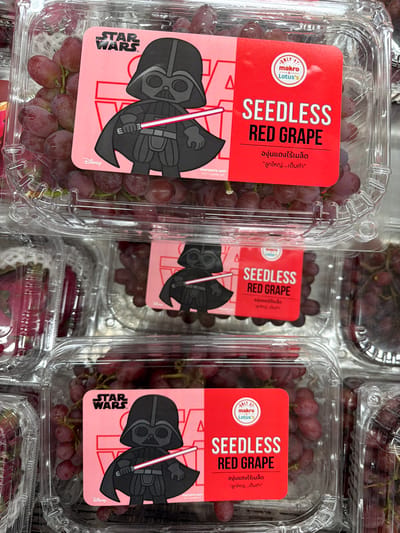 Three clear plastic containers of red seedless grapes are stacked vertically. Each container features a pink and red label with a cartoon illustration of Darth Vader from Star Wars. The top container's label shows "STAR WARS" at the top left, with Darth Vader holding a red lightsaber and the text "SEEDLESS RED GRAPE" in large white font. Below this, in Thai script, it reads "องุ่นแดงไร้เมล็ด" and "ลูกใหญ่...เต็มคำ". A "Disney" logo is present at the bottom left. The middle and bottom containers have identical labels. The middle container's label also includes "ONLY AT makro Lotus's" at the top right. The background is blurred, suggesting a retail environment, likely a supermarket aisle in Thep Krasatti, Thailand, given the Thai script on the labels and the presence of "makro Lotus's".