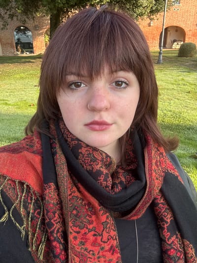 A medium shot captures a young adult female centrally positioned, looking directly forward with a neutral expression. She has medium-brown, shoulder-length hair with bangs covering her forehead, brown eyes, and fair skin displaying visible freckles across her nose and cheeks. A small silver hoop nose ring is present in her left nostril. Her lips are closed and have a light reddish tint. She wears a dark garment, partially obscured by a prominent scarf wrapped around her neck and shoulders. The scarf is primarily black with an intricate pattern of red, orange, and gold motifs, and features a tasseled fringe on the left side. A thin silver necklace is visible against her dark top.
The background consists of a sunlit, vibrant green lawn. Behind the lawn, to the left, stands a reddish-brown brick structure with a large arched opening reflecting the sky or distant scenery. To the right, a similar brick building with smaller arched features is visible, along with a black ornate lamppost and a sculpted green bush. The architecture and setting suggest a historical public space. This location is identified as a garden or courtyard area within the **Fortezza di Montepulciano** in Montepulciano, Italy.