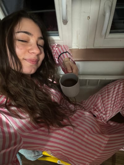 A woman in pink and white striped pajamas is lying down, holding a gray mug filled with liquid. She is near a white-framed window with a white radiator. Her brown, wavy hair frames her face. The background shows hints of other furniture and a blue towel. The image has a slightly tilted perspective.