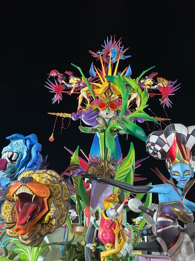 The image displays a collection of large, colorful, and intricately designed artistic figures, likely part of a festival or parade. The figures are made of various materials and are brightly painted, featuring a mix of human-like and animalistic elements.
In the foreground, a prominent sculpture depicts a fierce-looking lion's head with its mouth wide open, revealing a red tongue and sharp teeth. The lion's mane is rendered in yellow and brown spots. To the left of the lion, a blue, monstrous figure with large fangs and closed eyes is visible.
Dominating the center and upper portions of the image is a central figure with a regal and somewhat fantastical appearance. This figure wears large, circular red glasses, has green, leafy protrusions extending from its head and body, and is adorned with vibrant purple and blue elements. Surrounding this central figure are numerous sharp, spiky artistic elements in shades of red, blue, and yellow, resembling stylized flames or starbursts. Other smaller figures, some appearing human-like with stylized faces and colorful clothing, are integrated within the larger composition. One figure on the right has a blue face and wears a checkered jester's hat.
The overall scene is illuminated, suggesting it is either nighttime or a dimly lit indoor space, with the dark background emphasizing the vivid colors of the sculptures. The context of the image, particularly the style and scale of the figures, strongly suggests it is part of the "Fallas" festival in Alicante, Spain. This festival is known for its monumental satirical sculptures (ninots) that are erected in streets and plazas throughout the city and are traditionally burned on the final night of the festival. While the specific location within Alicante is not pinpointed in the image, the style of the figures is characteristic of Fallas displays. There are no discernible actions or interactions between people, as the focus is entirely on the static sculptures.