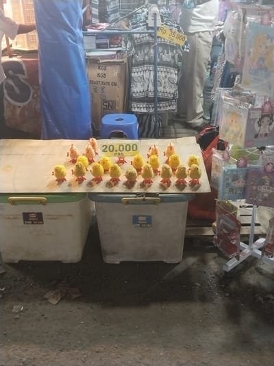 The image depicts an outdoor market stall in Sragen, Indonesia. A collection of small, wind-up toy chicks are arranged on a wooden plank resting on two plastic storage containers. A sign displays the price "20.000 PAS," presumably Indonesian Rupiah. Behind the stall, other merchandise is visible, including clothes, packaged toys, and a cardboard box with the dimensions "55x37,5x70 CM" labeled "KGS KGS" and an "SNI" label. A price tag on some clothing reads "Rp. 35.000 Pas." The ground appears to be a dark, paved surface with debris. People are visible around the stall.