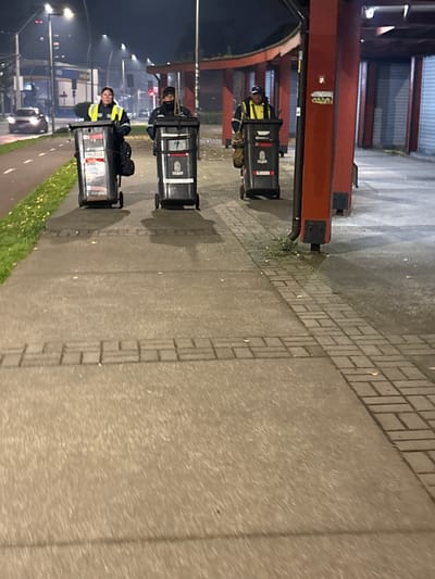 A street scene at night shows three workers in yellow safety vests riding small, motorized trash carts on a paved road with brick edging. Streetlights illuminate the road. Behind the workers is a bike path, a grassy area, and parked cars. A building structure with a red I-beam frame is visible on the left. There is no evidence to confirm this image was taken in the city of Osorno, Chile.