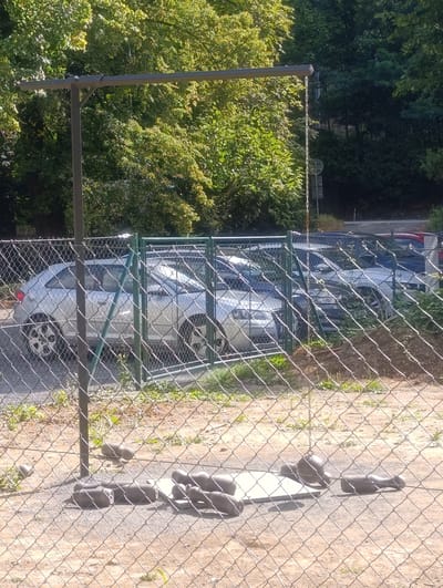 This image depicts an outdoor scene in Teplice nad Bečvou, Czechia, likely in a recreational or industrial area. A chain hangs from a horizontal bar supported by a vertical post on the left, suggesting a possible swing set or equipment for an outdoor activity. In the foreground, a wire mesh fence runs horizontally across the frame, obscuring a ground area covered with gravel and scattered with various heavy objects, possibly weights or parts of outdoor exercise equipment. Beyond this fence, another wire mesh fence encloses a parking lot containing several silver and dark-colored cars. The background is dominated by lush green trees, indicating a natural or park-like setting. The overall impression is one of a functional, possibly disused, outdoor space with elements of both recreation and utility.