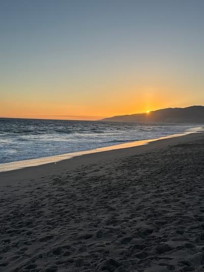 The image depicts a serene sunset scene at a beach in Malibu, California. The sky transitions from a pale blue at the top to a warm orange glow near the horizon, where the sun is partially obscured by a distant, silhouetted mountain range. The ocean stretches across the middle ground, with waves gently rolling towards the shore. The water reflects the golden hues of the sunset, creating a shimmering path across its surface. The foreground is dominated by dark, sandy beach, marked with numerous footprints, suggesting recent human activity. The overall atmosphere is tranquil and picturesque.