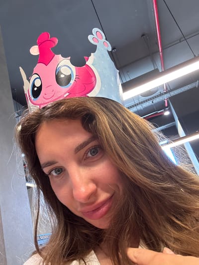 A tilted close-up shot shows a woman with light brown hair and blue-green eyes, posing next to a cartoon drawing of Pinkie Pie from My Little Pony. The background is an out-of-focus industrial-style ceiling with exposed pipes and fluorescent lighting. The location is in the city of Sueca, Spain.