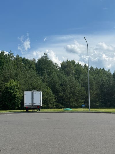 A white box truck with the license plate AB392B-7 is parked on an asphalt surface in front of a dense forest of green trees. A tall, dark lamp post stands to the right of the truck. In the background, a bright blue sky is visible with scattered white clouds. There are no people visible in the image.