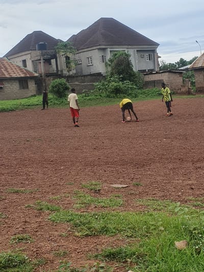 The image depicts an outdoor scene in Idah, Nigeria, featuring four young men on a reddish-brown dirt and gravel field with scattered patches of green grass. In the mid-ground, a man in a yellow and black patterned long-sleeved shirt and dark shorts bends over a white and black soccer ball, wearing pink athletic shoes. Another man, similarly dressed in a yellow and black patterned shirt and dark shorts with orange athletic shoes, stands upright to his right. Further left, a man in a light t-shirt and red shorts stands with his back to the viewer, and a fourth man in a dark tank top and dark shorts stands to the far left. The background consists of residential buildings under an overcast sky. On the left, a single-story building with a rusted corrugated metal roof and concrete walls is visible. In the center, a prominent two-story concrete building with a dark, multi-hip roof, multiple white-framed windows, and a black water tank on a smaller adjacent roof structure is present. To the right, another single-story building with a rusted roof and a distant street light pole are visible. Green vegetation, including bushes and trees, grows around the buildings and along the edges of the field.