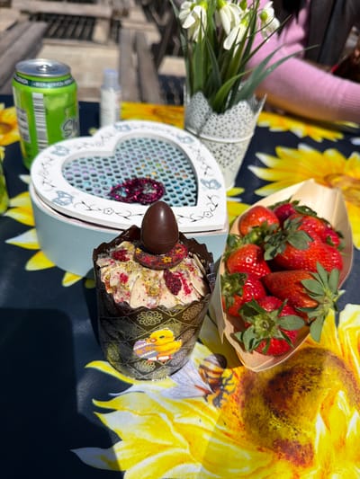 A bright and sunny outdoor scene captures a festive arrangement of treats and decorative items laid out on a table. The table is covered with a dark blue/black tablecloth featuring a vibrant pattern of large yellow sunflowers and bees.
In the foreground, a decorative cupcake or muffin is prominently displayed. It's encased in a dark brown and gold patterned paper wrapper, adorned with a yellow Easter chick sticker. The cupcake is topped with a generous swirl of light-colored frosting, sprinkled with red dried fruit pieces (possibly raspberries) and green pistachios. A small, dark chocolate egg rests on top, accompanied by a piece of decorated chocolate.
To the left, a heart-shaped light blue or grey decorative box is visible. Its front panel has a mesh-like design, revealing a glittery red heart inside. The box edges are decorated with delicate white floral and butterfly patterns. Next to it, a small bouquet of white flowers, resembling snowdrops, is wrapped in white paper with a lace-like texture.
Further back, a small wooden boat-shaped container is filled with fresh, ripe red strawberries, their green stems providing a vibrant contrast. Partially visible on the left are a green beverage can with a barcode and some text, possibly "WEIHSS... BIERE...", and a small clear plastic bottle. A person wearing a pink sleeve is subtly visible in the upper left corner, suggesting human presence.
The bright daylight indicates a clear, sunny day, creating a cheerful and celebratory atmosphere, possibly for an Easter gathering or picnic in an outdoor setting like Monistrol de Montserrat, Spain.