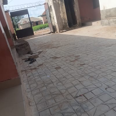 The image depicts an outdoor scene in Wukari, Nigeria, featuring a paved walkway in front of a building with a gate. The walkway is constructed of patterned concrete interlocking pavers. To the left, a building wall painted in a reddish-brown hue is visible. A large, dark metal gate, partially open, is situated at the entrance, topped with barbed wire. Beyond the gate, residential buildings with corrugated metal roofs and surrounding vegetation can be seen. In the foreground, on the paved walkway, a pair of black flip-flops are placed. The ground shows some areas of wear and tear on the pavers, with some dirt and debris scattered about. The overall impression is of a somewhat aged urban or semi-urban environment.