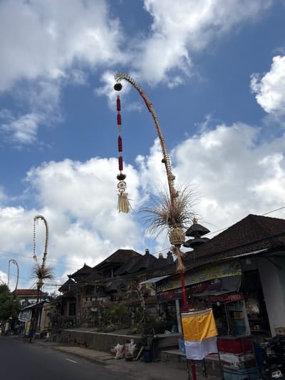 In Tampaksiring, Indonesia, a low-angle, full shot captures a street scene under a partly cloudy blue sky. Two elaborately decorated tall, curved poles, known as Penjor, stand prominently in the foreground, adorned with red flower garlands and natural materials like straw and dried leaves. One Penjor is positioned to the left, its curved form reaching towards the sky, while another, larger Penjor dominates the center-right of the frame, its intricate top section featuring woven elements and a feathery, grass-like decoration.
Beneath the Penjor, traditional Balinese architecture is visible, characterized by dark, tiled, multi-tiered roofs. The buildings appear to be shops or residences, with signs in Indonesian text, some displaying prices in Rupiah (RS). Evidence of commerce is present, with items like bottles, containers, and bundled goods displayed outside. A few individuals are visible on the street, including a person walking on the left and what appears to be a cart. The road in the foreground is asphalt. The overall impression is one of a vibrant, culturally rich street in an Indonesian town.