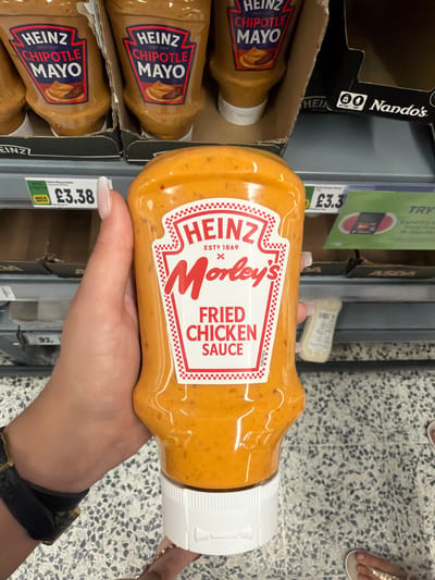 This is a close-up, vertical shot of a person's hand holding a bottle of Heinz Morley's Fried Chicken Sauce in a supermarket aisle. The sauce bottle is orange and has a red and white label with the Heinz logo and "Morley's Fried Chicken Sauce" written on it. To the left of the sauce bottle are two bottles of Heinz Chipotle Mayo, also in cardboard packaging. A price tag of £3.38 is visible below the Chipotle Mayo bottles. In the background, shelves with various products and a section of speckled flooring are visible. The lighting suggests an indoor setting, typical of a supermarket. The image appears to have been taken during the day. There is no indication of the weather or specific time of day.