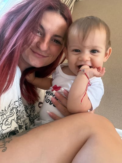 A woman with red hair and a baby are posing for a selfie. The woman is on the left, and the baby is on the right, being held by the woman. The baby is wearing a white onesie with red lettering that reads "everything is love." The baby has a red string bracelet on its wrist. The background appears to be a wall with vertical blinds or drapes. The image does not contain any identifiable landmarks or context to determine its location within the city of Veghel, Netherlands.