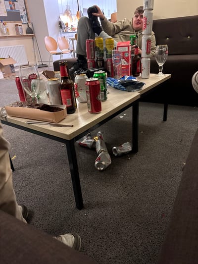 The image shows a living room in Lancaster, UK (latitude: 54.007, longitude: -2.786). A young man sits on a brown sofa, wearing a white shirt and black pants. His legs are crossed, and he is holding a beer bottle in his right hand. A stack of six beer cans stands on the table in front of him, and there are several other beer cans scattered around the table. There is a glass of beer on the table next to him. A piece of chocolate cake is sitting on the table, along with a pair of glasses. There are several empty beer cans on the floor beneath the table, and several empty cans on the floor. A pair of brown chairs are situated in the background, with one being used by the man. The room has gray carpet. The man is looking off camera. He is the only person in the scene.